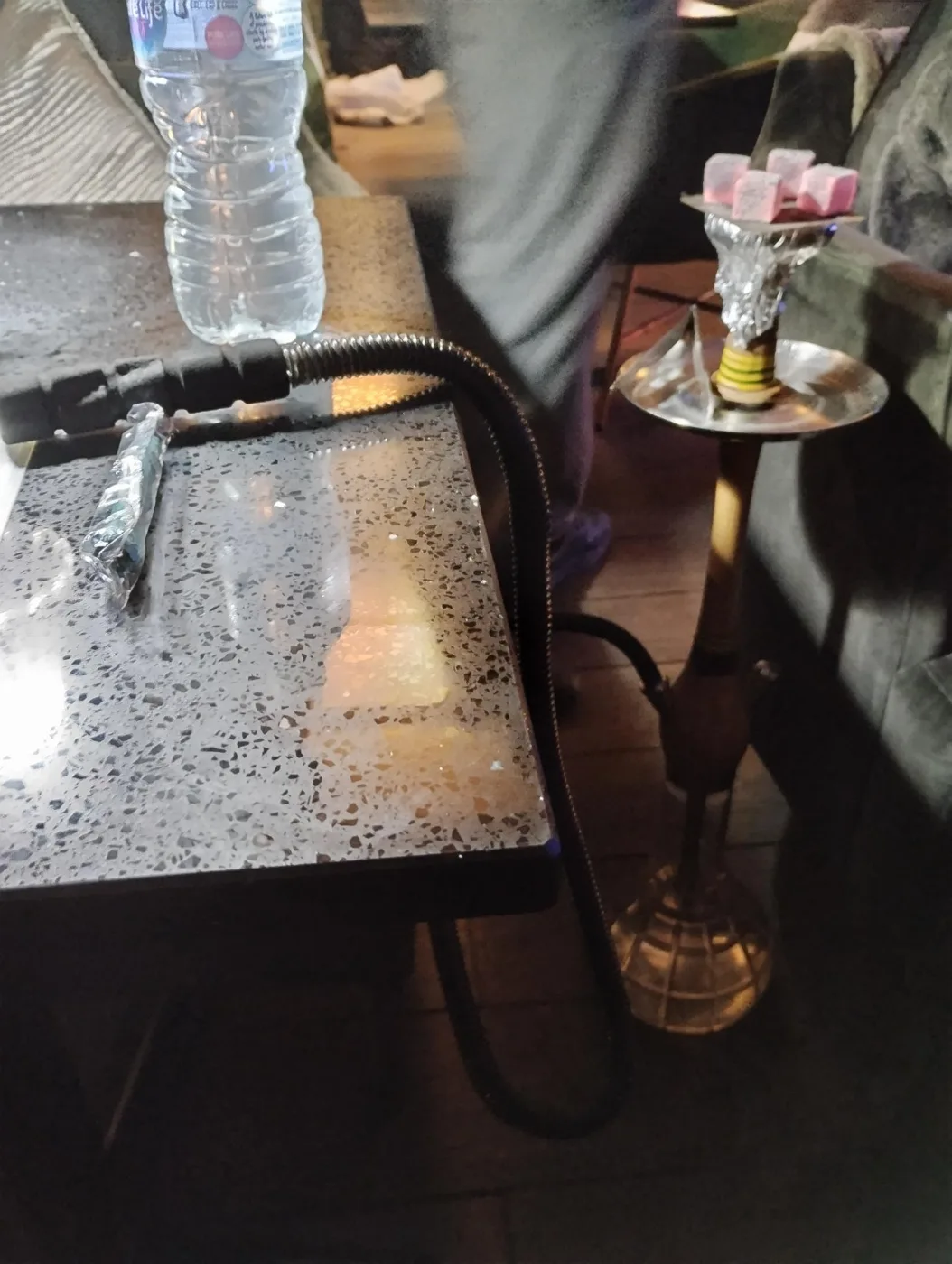 A shisha pipe with lit coals on a foil-wrapped bowl beside a speckled stone table, with a hose and a bottle of water on the table