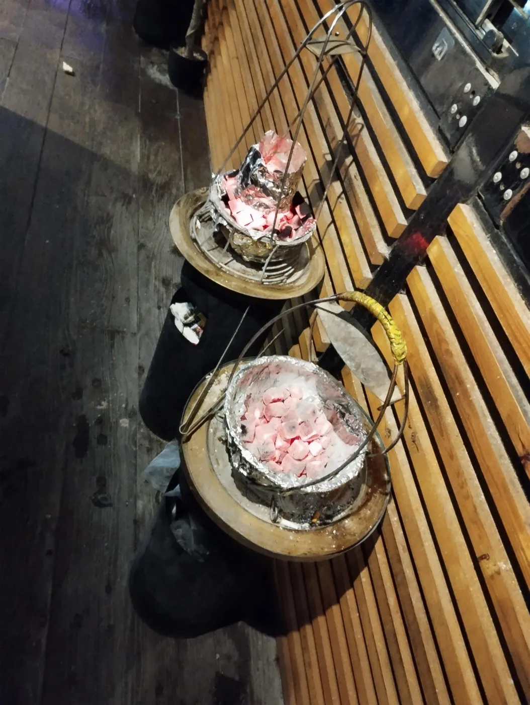 Two metal baskets of glowing charcoal in foil on stands beside a slatted wooden wall