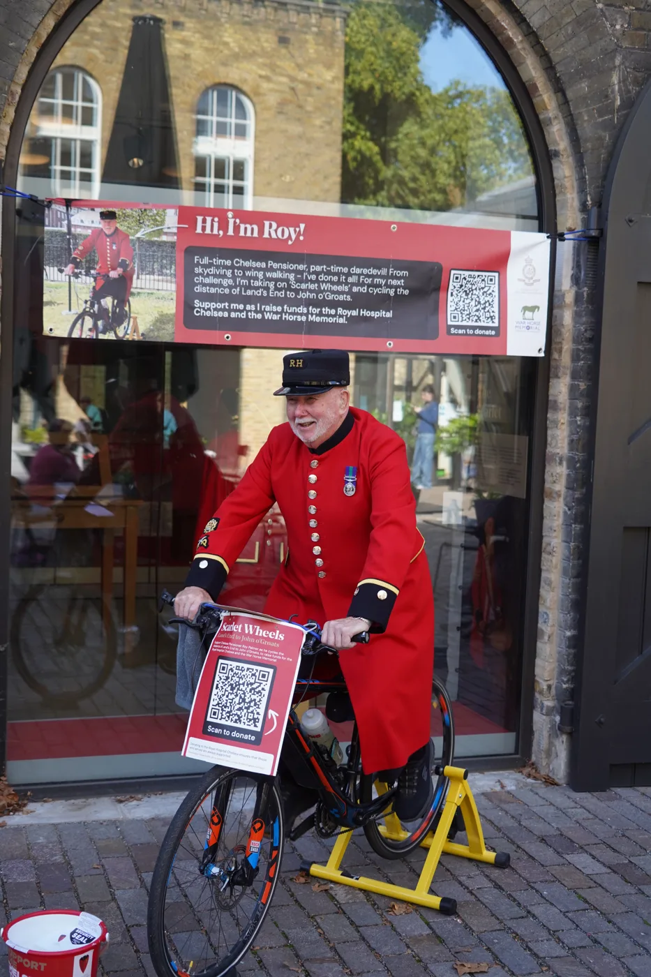 Roy, a Chelsea Pensioner in a scarlet coat and black RH cap, smiles on a bicycle fixed to a yellow static trainer in front of an arched glass doorway, under a red banner reading Hi, I'm Roy!, with a red collection bucket beside him