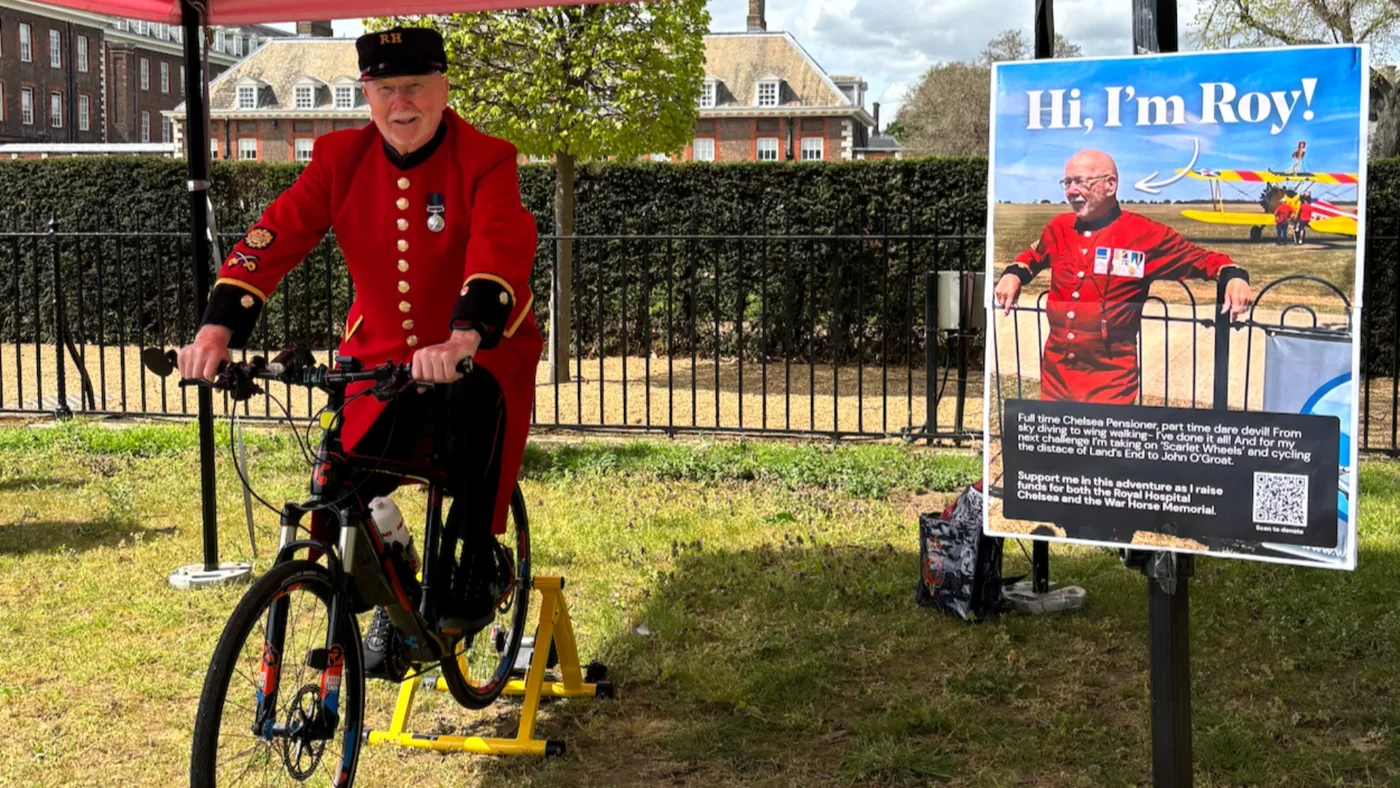 Roy in his scarlet coat on a bike on a yellow static trainer under a red gazebo on grass, with railings and the red-brick Royal Hospital buildings behind, beside a Hi, I'm Roy! poster showing him in front of a yellow biplane