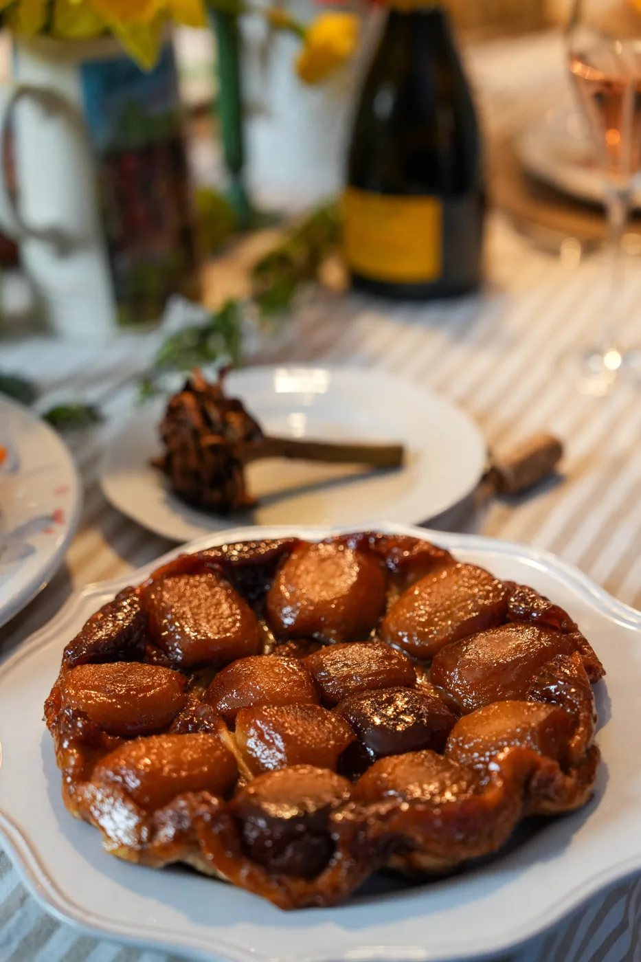 A caramelised apple tart on a white plate on a table with a striped cloth, a bottle and glasses behind