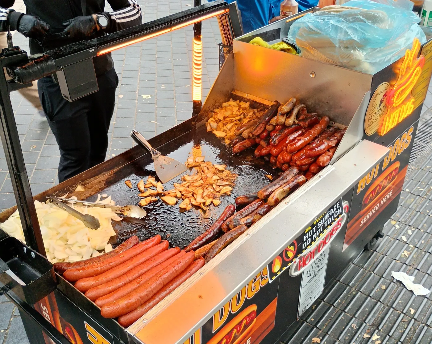 A hot dog cart with sausages and onions on its hotplate, standing on a paved street