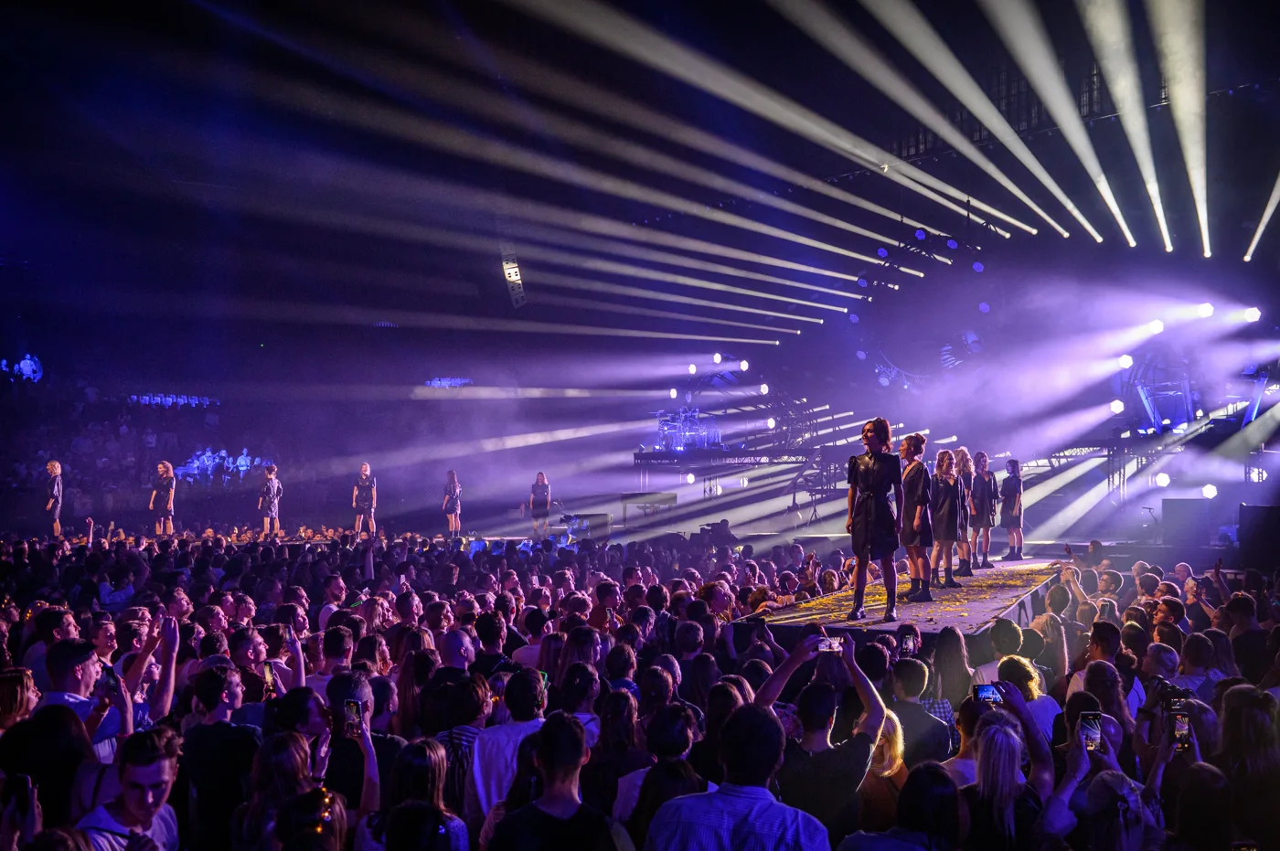 Scala performing on a long lit stage in front of a packed standing crowd, with beams of light overhead